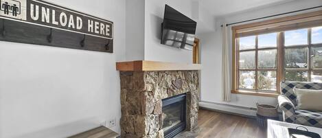 A living room with a stone fireplace, a mounted television, and a large window with a view of a snowy landscape.