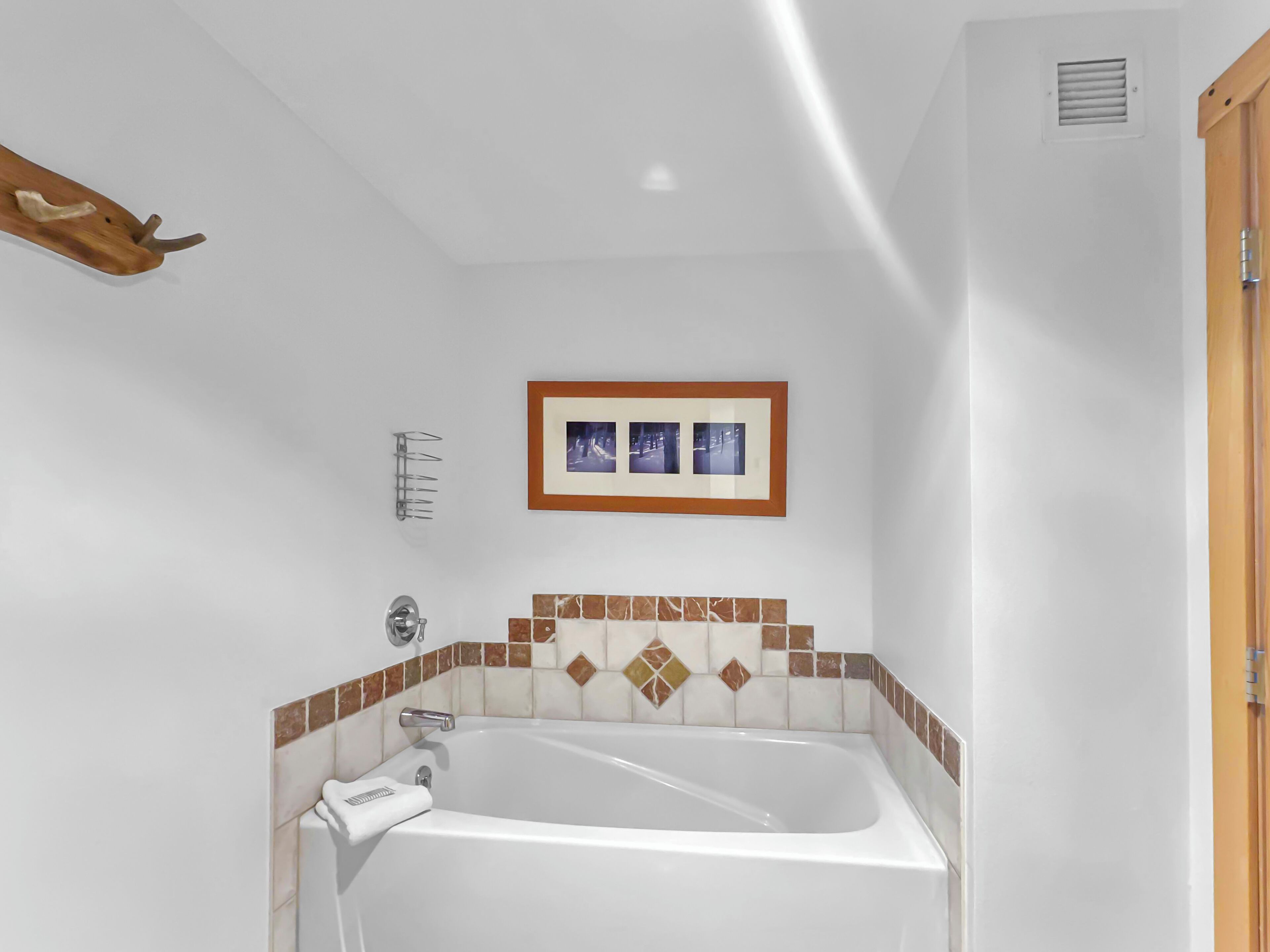A bathroom with a white bathtub, a wooden towel rack, a framed picture on the wall, and a tiled shower area.