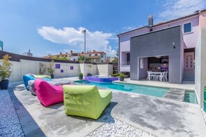 Un área de piscina moderna al aire libre con sillones tipo sacos de frijoles de colores, una piscina pequeña y un patio con mesa y sillas.