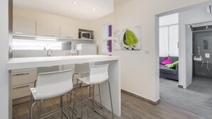 A modern kitchen with white cabinetry, a white countertop, and a microwave. There are two white bar stools and a painting of a green leaf on the wall.