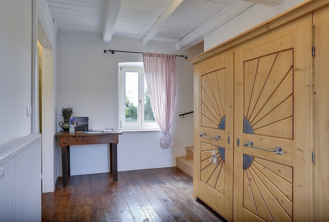A hallway with wooden flooring, a wooden wardrobe with two doors, a window with a curtain, and a small table with a plant and a book.