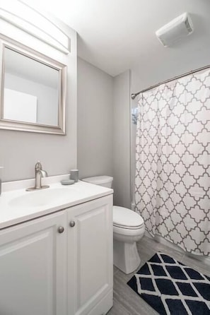 Une salle de bain avec un meuble-lavabo blanc, des toilettes, un rideau de douche à motifs géométriques et un tapis noir et blanc.