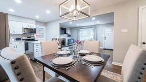Un coin repas avec une table dressée pour quatre personnes, un lustre et une cuisine en arrière-plan.