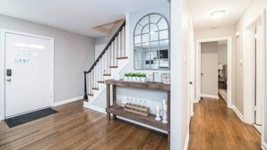 Un couloir doté d’un plancher en bois, d’une porte blanche, d’un escalier avec une rampe noire et d’une étagère ornée d’objets décoratifs.