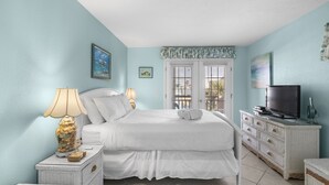 A bedroom with a bed, bedside tables, a TV, and a window with blinds. - Sunset Terrace 201 · Gulf Front · Gulf Front (Bradenton Beach)