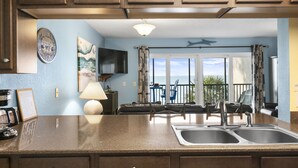 A kitchen with a large island, double sink, and wooden cabinets. There is a dining area with chairs and a table, and a balcony with a view of the ocean. - Sunset Terrace 201 · Gulf Front · Gulf Front (Bradenton Beach)