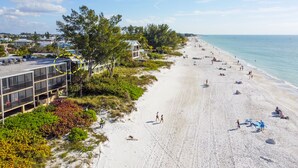 A beach with a building featuring large windows and balconies, surrounded by greenery and a clear sky. - Sunset Terrace 201 · Gulf Front · Gulf Front (Bradenton Beach)