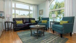 A living room with a dark leather sofa, teal armchairs, a round coffee table, and a large window with curtains. - Dolphin Dance South · Gulf Views · 3-Night Min (Bradenton Beach)