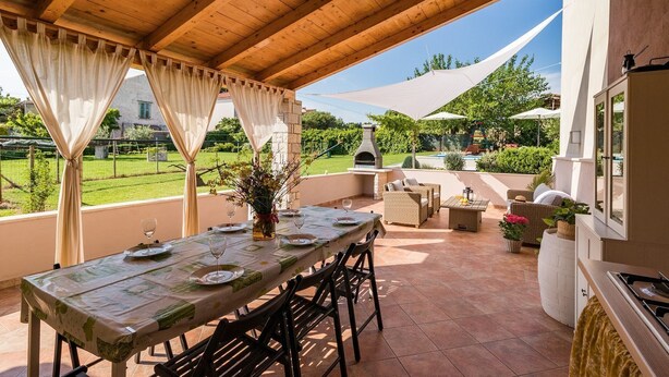 An outdoor patio with a dining table set for four, surrounded by wicker furniture and a stone oven.