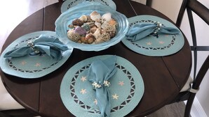 A round dining table with a white cushioned bench, surrounded by four chairs. The table is set with a blue woven placemat, a bowl of decorative seashells, and a blue napkin. - 100 PLUS 5 STARS! BEACHFRONT! FREE BEACH CHAIRS! STAND UP SHOWER! FULLY STOCKED! (Panama City Beach)