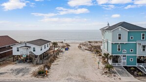 A beachfront property with a sandy path leading to the ocean, a white building with a balcony, and a larger teal building with a sign. - Family oasis beach retreat in quiet woodlands (Oak Island)