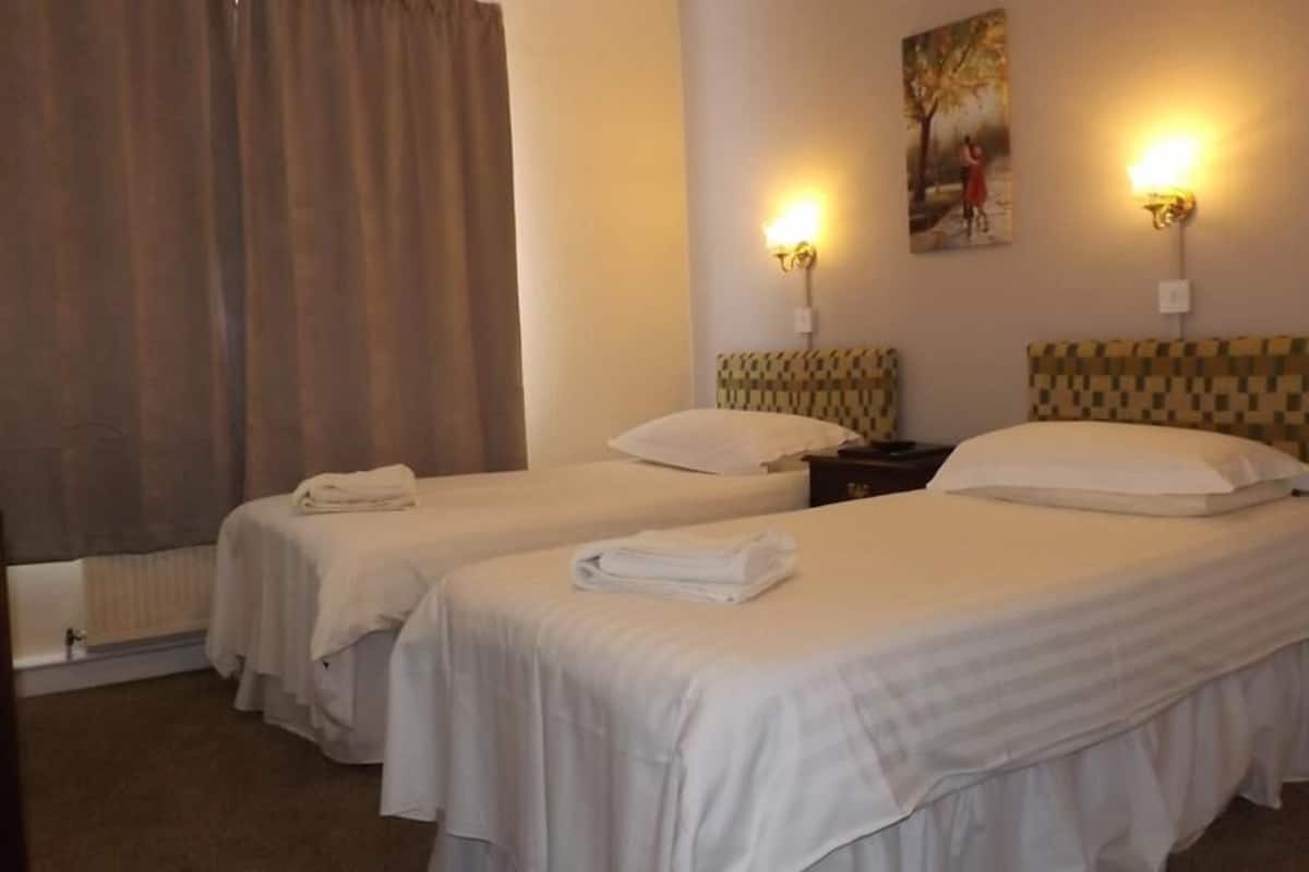 A hotel room with two beds, each with a pillow and a folded towel. There are wall-mounted lights and a painting above the beds.