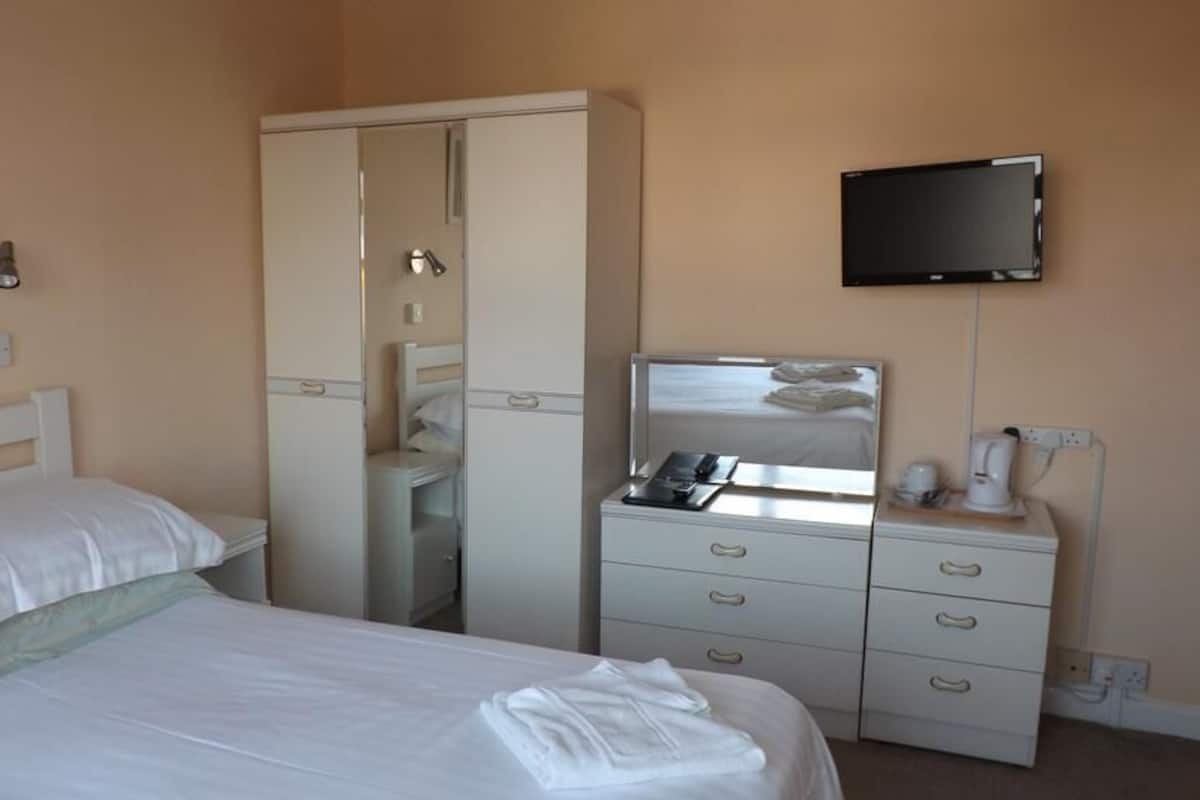 A hotel room with a bed, a wardrobe, a desk with a flat-screen TV, and a bedside table with a lamp and a phone.