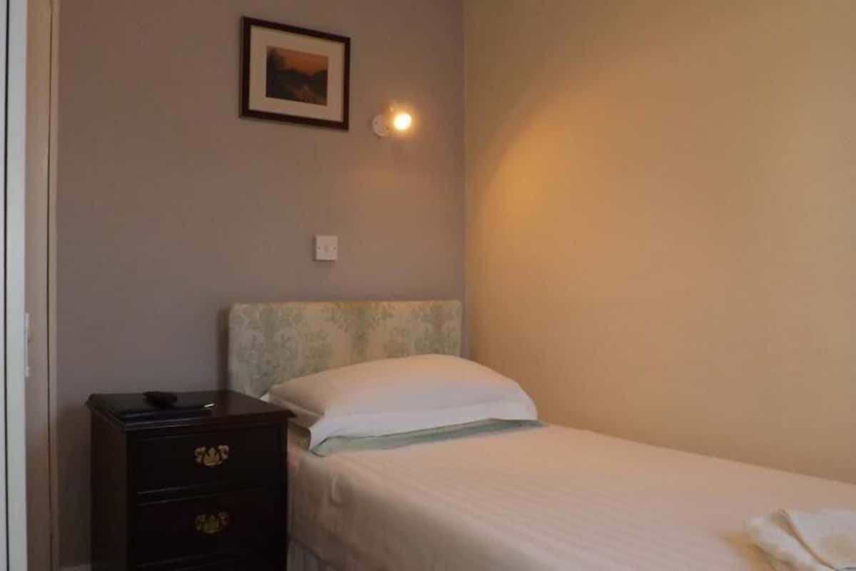 A small, simple room with a bed, a nightstand, and a wall-mounted light.