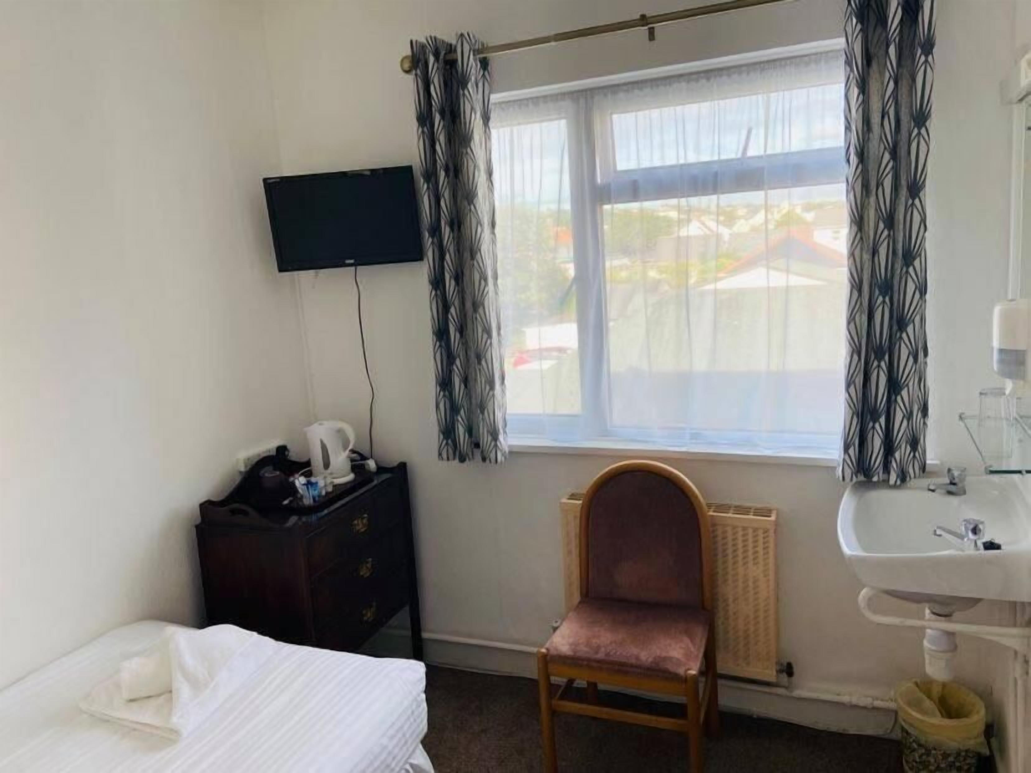 A hotel room with a bed, a desk with a kettle, a chair, a TV, a window with curtains, and a bathroom with a sink.