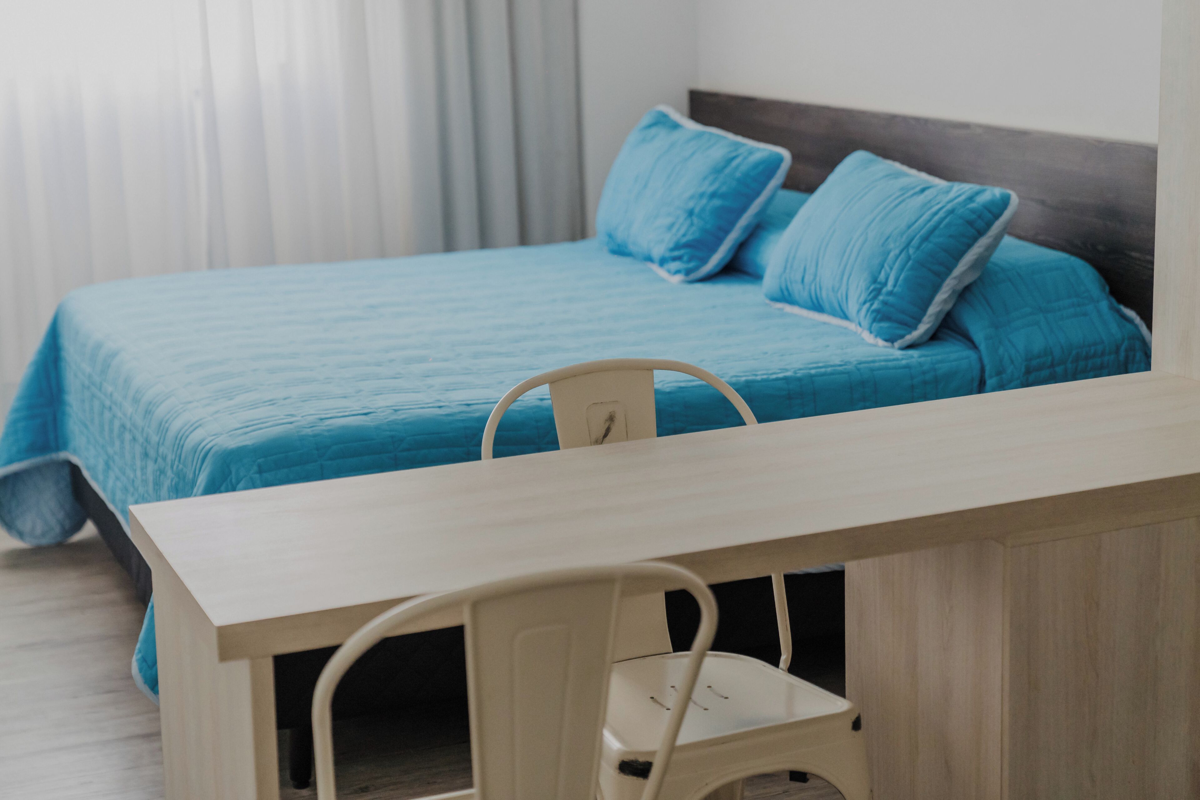 Una cama con colcha y almohadas azules, una mesita de noche de madera y una silla blanca.