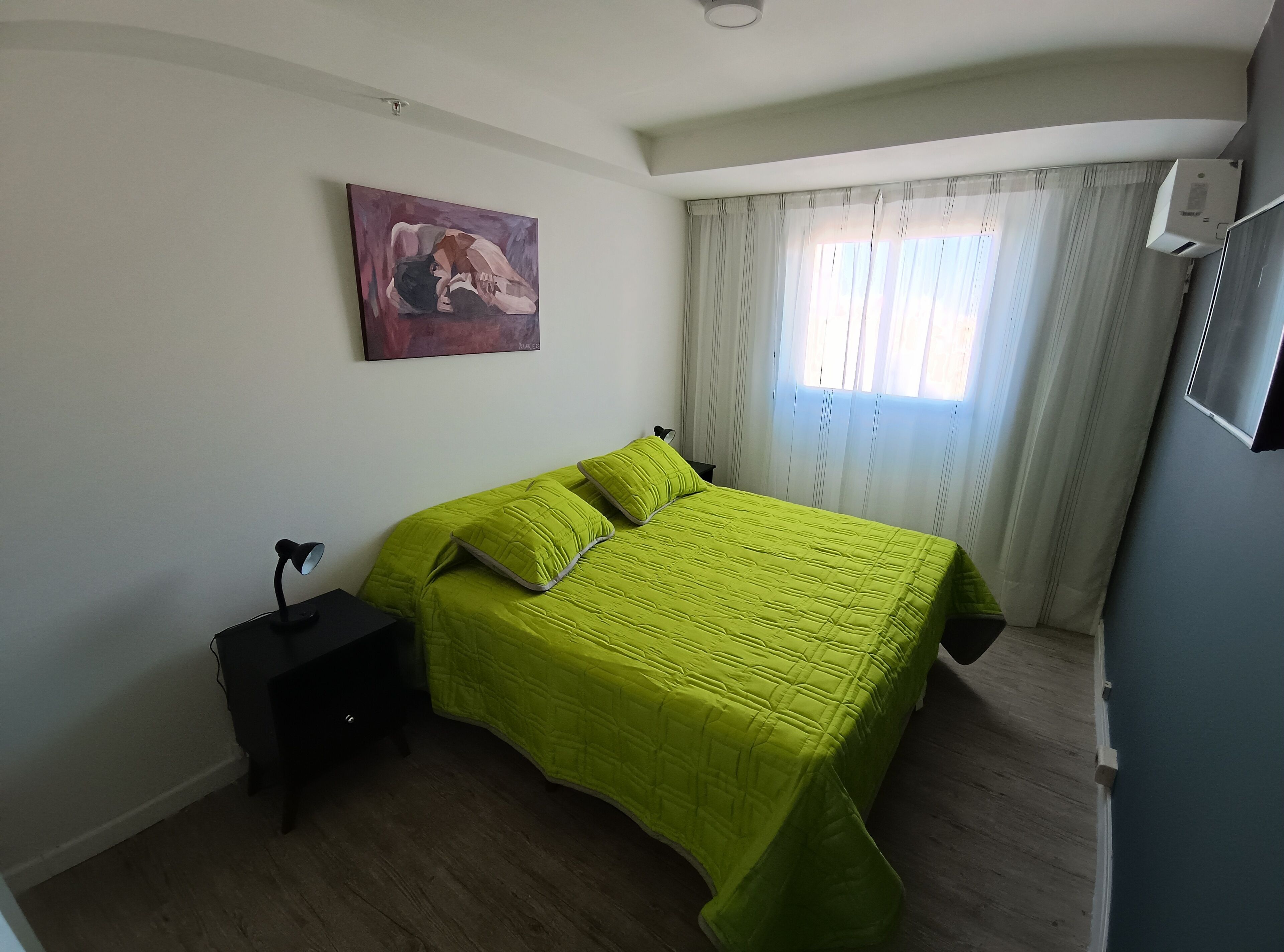 Un dormitorio con cama, mesita de noche, lámpara, un cuadro y ventana con cortinas.