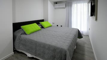 A neatly arranged bedroom with a bed, pillows, an air conditioning unit, a bedside table, and a window with curtains.