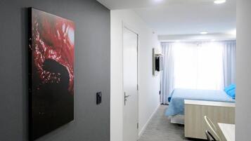 A modern hotel room with a grey wall, a large abstract painting, a wooden floor, a bed with blue bedding, and a desk with a chair.