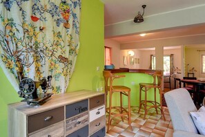 Una habitación con una pared verde, un armario de madera con cajones, dos taburetes de bar de madera, un sofá blanco y una cortina estampada.