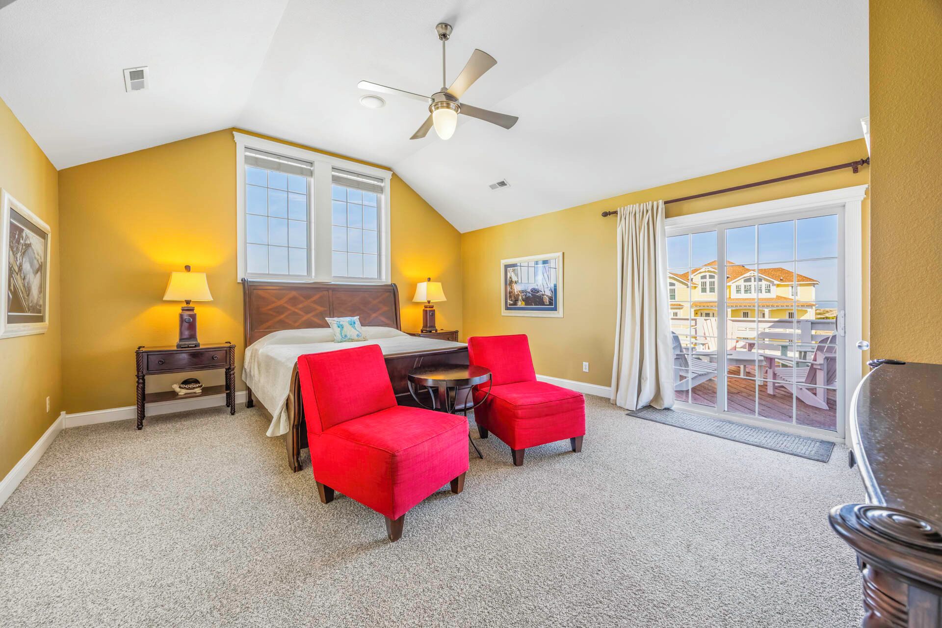 A hotel room with a bed, two red chairs, a table, a ceiling fan, and a balcony with outdoor furniture.