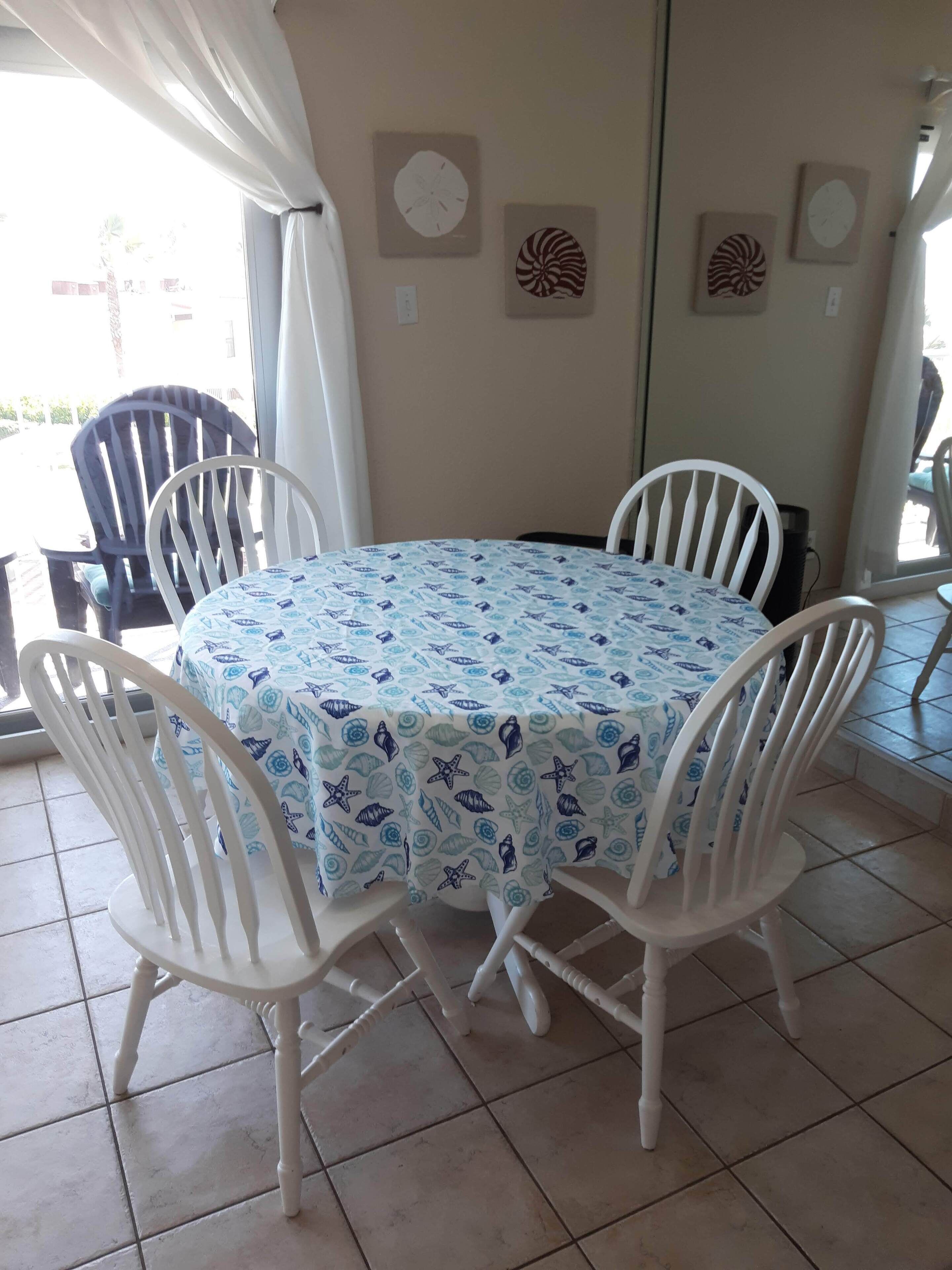 Un coin repas avec une table ronde recouverte d’une nappe bleue et blanche à motifs, entourée de chaises blanches.