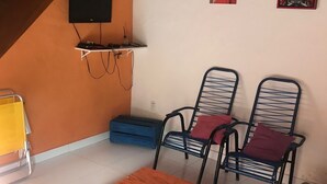 Un salon avec une table basse en bois, deux chaises en métal avec des coussins, un téléviseur fixé au mur et un escalier.