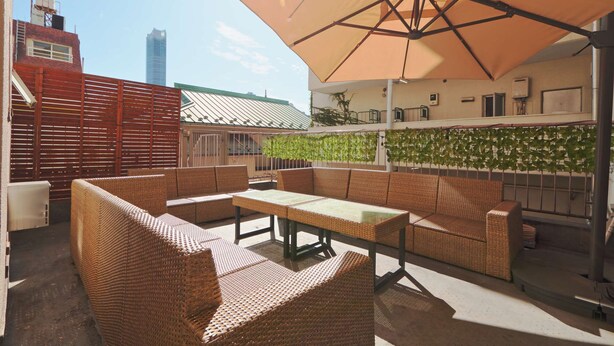 Un patio al aire libre con mobiliario de mimbre, un paraguas grande y vista a un edificio con techo verde.