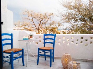 Una terraza con dos sillas azules, una mesita pequeña, un jarrón decorativo y una pared con ventanas cuadradas.