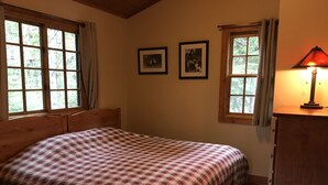 A bedroom with a wooden bed, two framed pictures on the wall, a window with curtains, and a nightstand with a lamp. - Doig Cabin: Support a great cause while on vacation! (Government Camp)