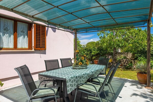 An outdoor patio with a table and chairs, covered by a blue mesh canopy, and surrounded by greenery. - Fully fenced holiday home, your pet is also welcome (Rakalj)