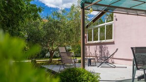 A covered outdoor seating area with chairs and a table, surrounded by greenery and a building with large windows. - Fully fenced holiday home, your pet is also welcome (Rakalj)