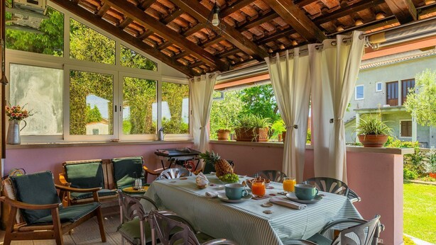 A covered outdoor dining area with a table set for two, surrounded by chairs, potted plants, and a view of greenery and buildings. - Fully fenced holiday home, your pet is also welcome (Rakalj)