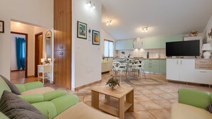 A modern living room with green sofas, a wooden coffee table, and a dining area with white chairs. A kitchen with light green cabinets and a black TV is visible in the background. - Fully fenced holiday home, your pet is also welcome (Rakalj)