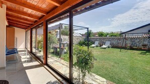 Une terrasse couverte, dotée de grandes fenêtres donnant sur une cour verdoyante et un mur en pierre.