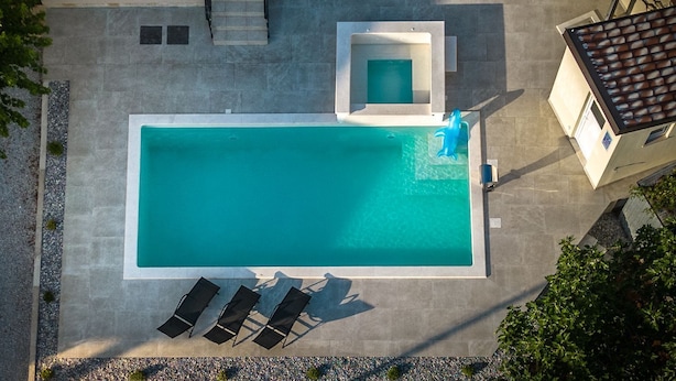 An aerial view of a rectangular swimming pool with a smaller adjacent pool, surrounded by lounge chairs and a staircase leading to an upper level.