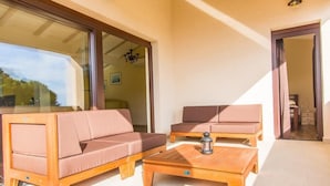 A sunlit room with two sofas and a wooden coffee table, large glass doors leading to a balcony, and a chandelier visible through the glass.