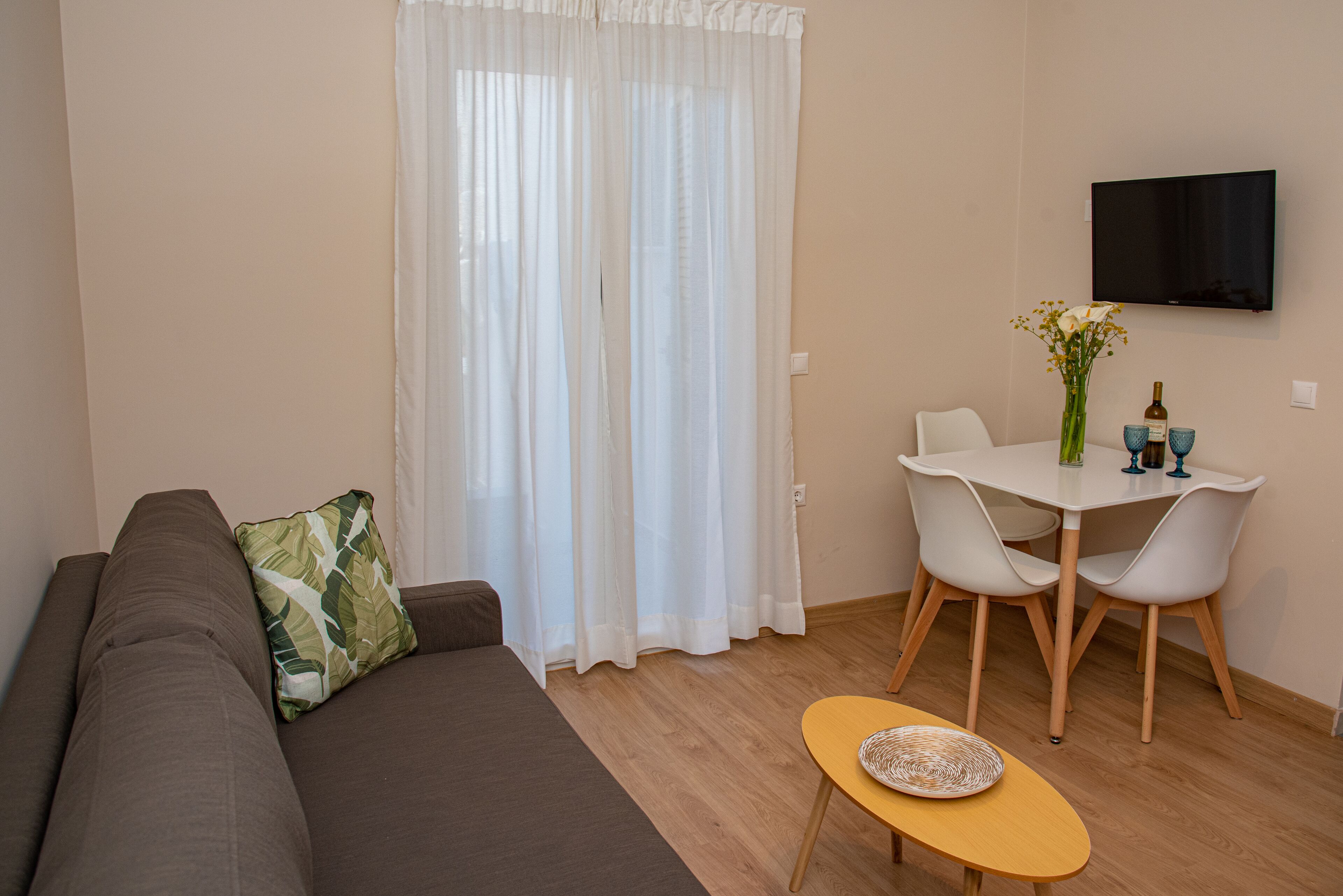 Un salon comprenant un canapé, une petite table avec des chaises, un téléviseur fixé au mur et une fenêtre ornée de rideaux légers.