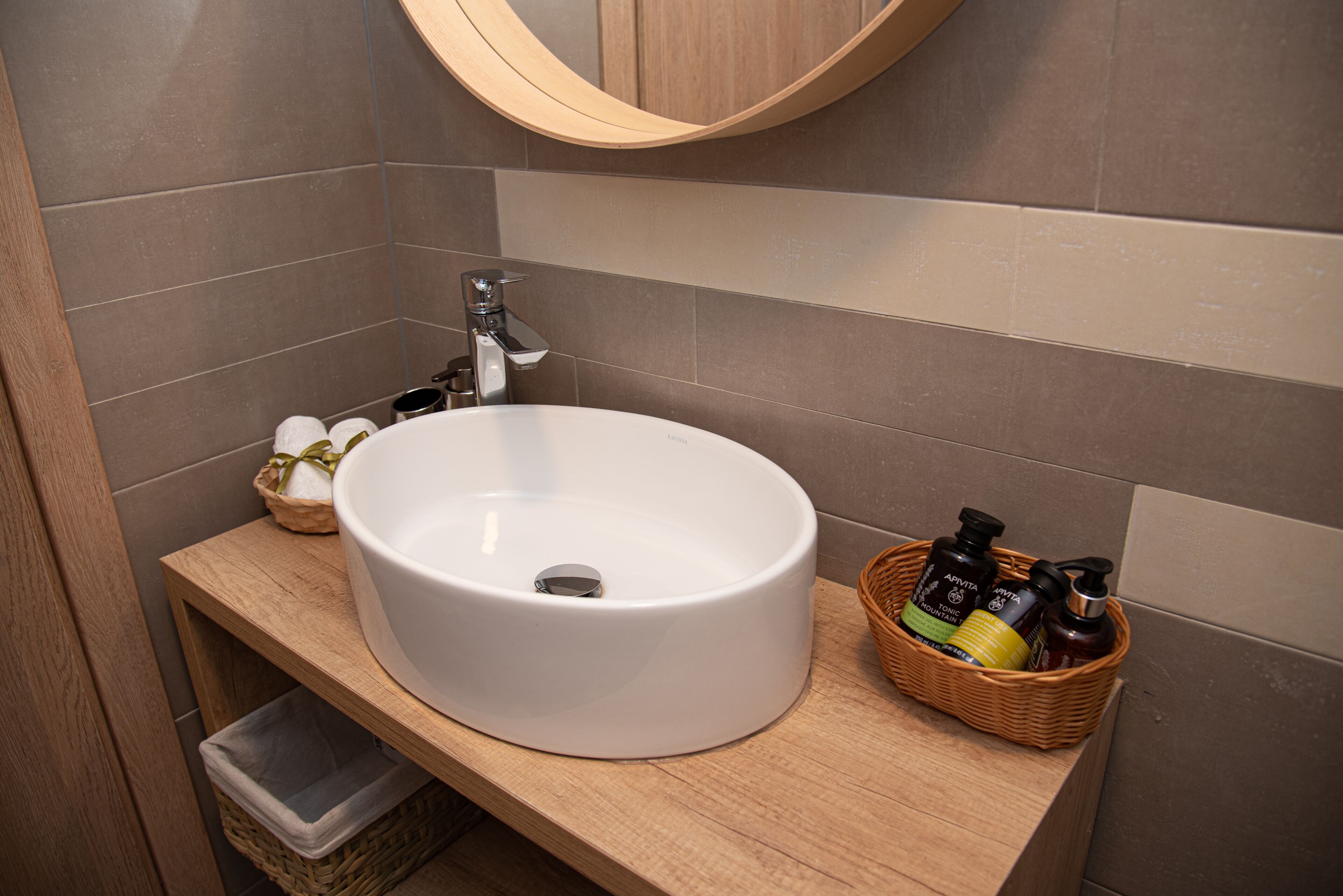 Une salle de bain avec un lavabo blanc, un meuble-lavabo en bois, un panier en osier contenant des articles de toilette et une poubelle.