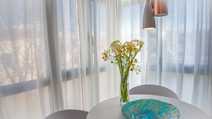 Una mesa con un jarrón de flores y un plato decorativo, frente a cortinas translúcidas y vistas al exterior.