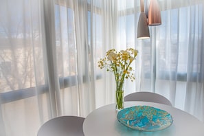 A table with a vase of flowers and a decorative plate, set against a backdrop of sheer curtains and a view of the outdoors.