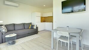 Een moderne woonkamer met een grijze bank, een witte eettafel met stoelen, een flatscreen-televisie aan de muur en een keuken in de achtergrond.