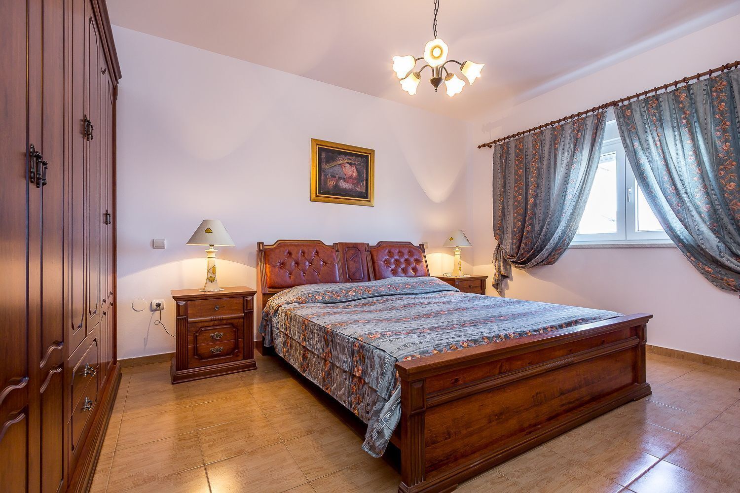 Een slaapkamer met een houten bed, twee nachtkastjes, een kaptafel, een kroonluchter, een raam met gordijnen en een kledingkast.