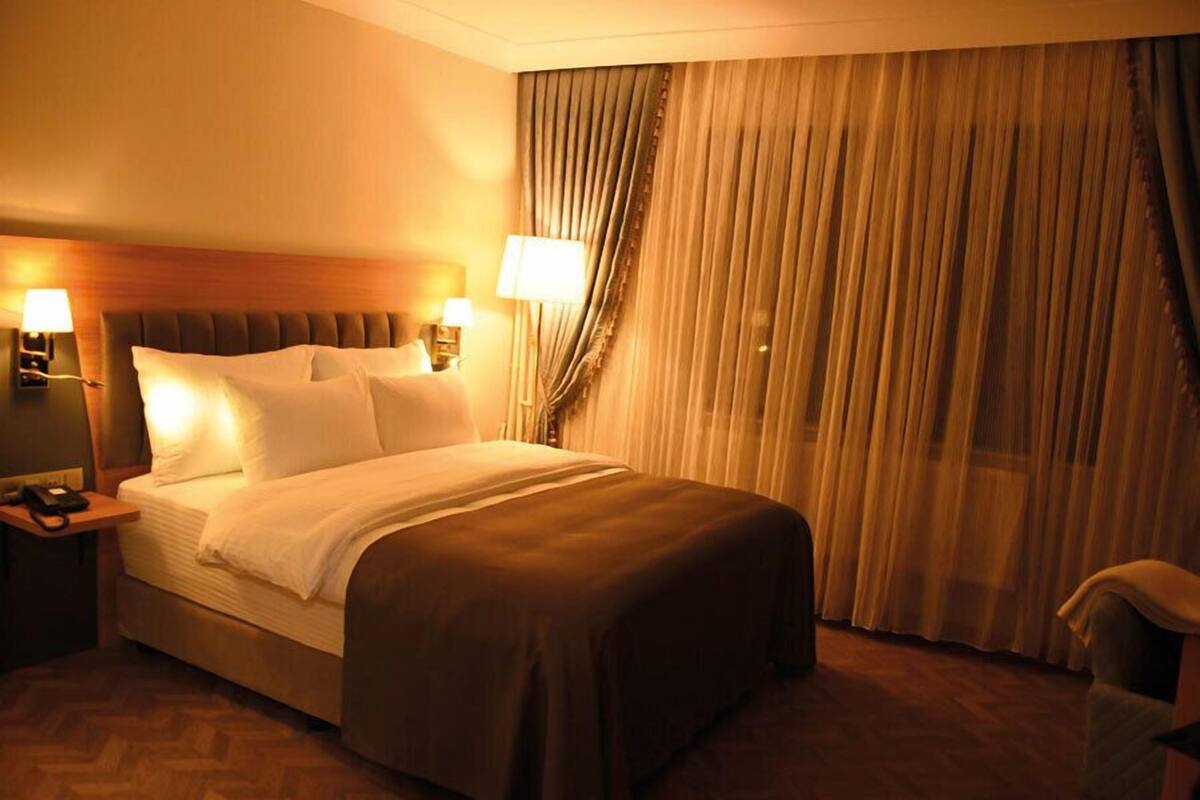 Una habitación de hotel con una cama grande, dos lámparas de noche, una mesita de noche y una cortina.