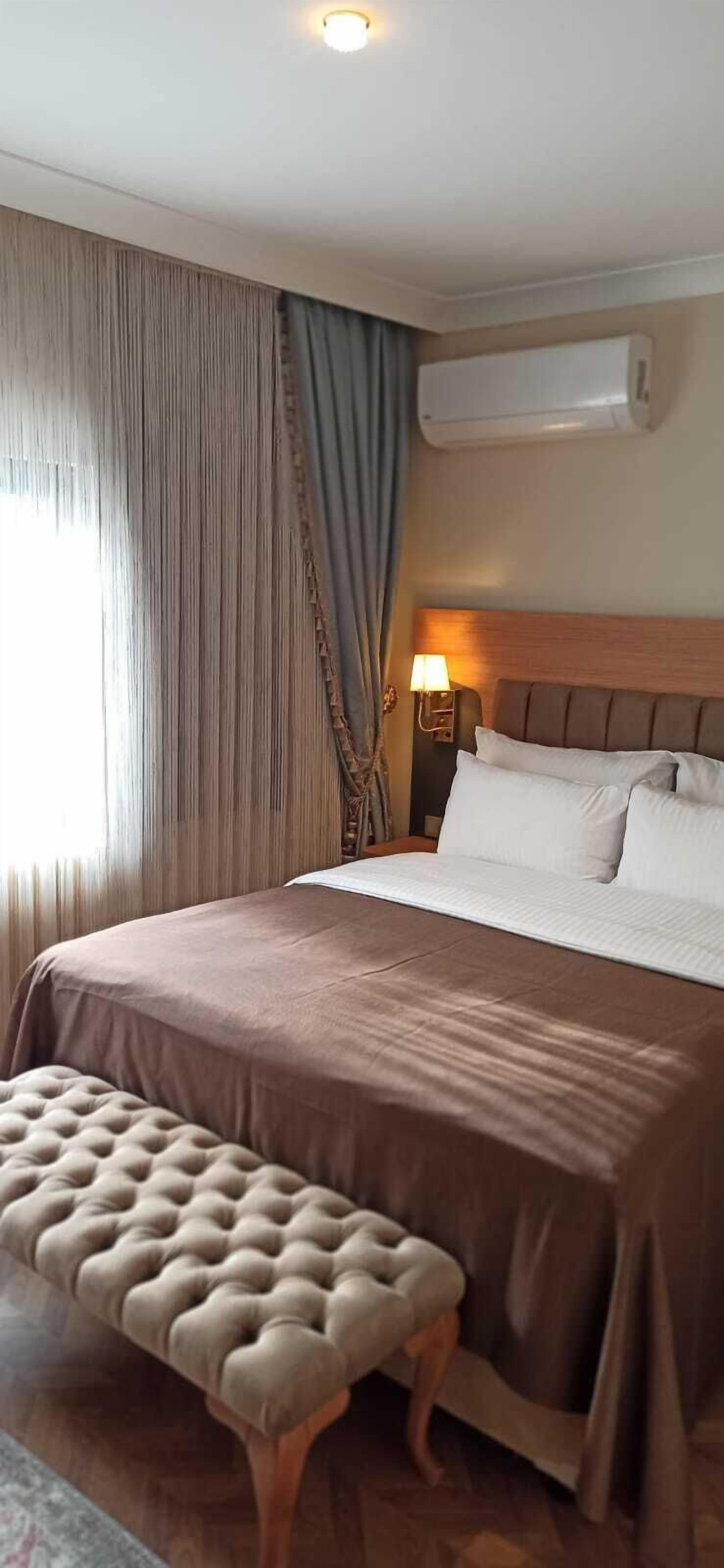 Habitació d'hotel amb un llit gran, una taula de nit amb una làmpada, una banca i un aparell d'aire condicionat.