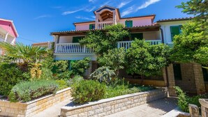 A two-story building with a balcony, surrounded by lush greenery and a stone retaining wall.