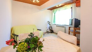 A room with a bed, a chair, a small table, a radiator, a television, and a window with curtains.