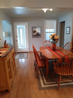 A dining area with a wooden table, chairs, and a vase of flowers. There is a door leading to another room and a window with a view.