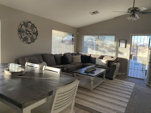 A modern living room with a sectional sofa, a dining table, and a coffee table. There is a large wall clock and a ceiling fan.
