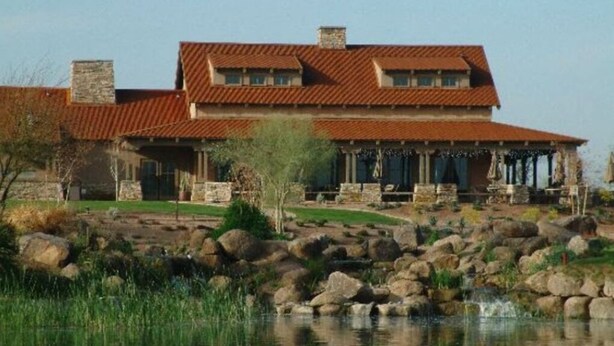A large, two-story house with a red-tiled roof, a stone chimney, and a stone porch with columns. In front of the house is a rocky landscape with a small pond and a waterfall. - *** GORGEOUS DESERT GETAWAY WITH HEATED  POOL NEAR THE DUKE GOLF COURSE*** (Maricopa)